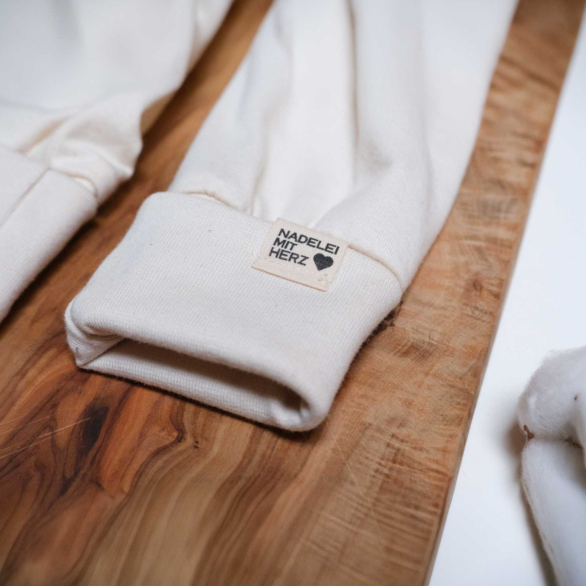 Nahaufnahme des Beinbündchens einer naturfarbenen Abhaltehose in Gr. 56. Zu sehen ist das Mitwachsbündchen, am Bündchen ist ein Label mit „Nadelei mit Herz“ angebracht. Die Hose liegt auf Holz, dieses wiederum auf einem hellen Hintergrund.
