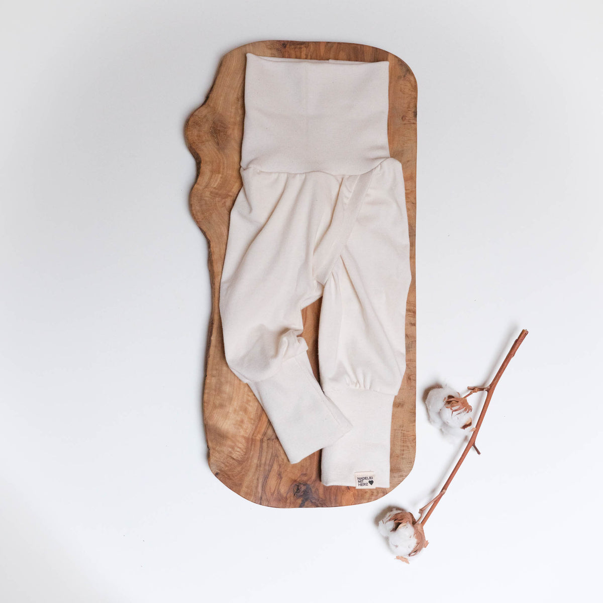 Abhaltehose in Größe 56, Farbe natur, mit ausgeklappten Mitwachsbündchen, auf Holz, mit einem Baumwollzweig.